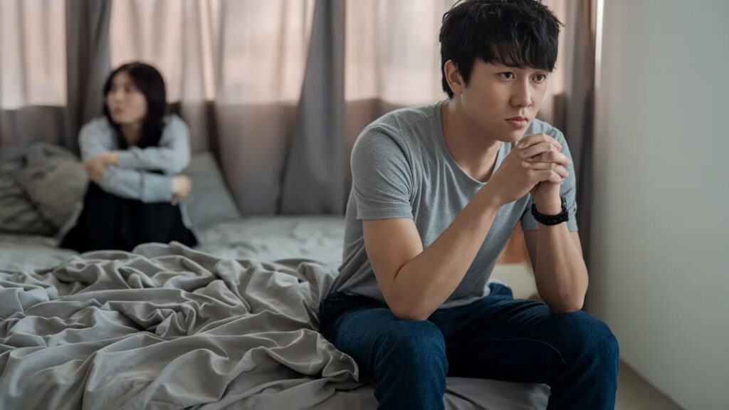 年輕情侶在房間中，男子憂心忡忡地坐在床上，女子則坐在床上