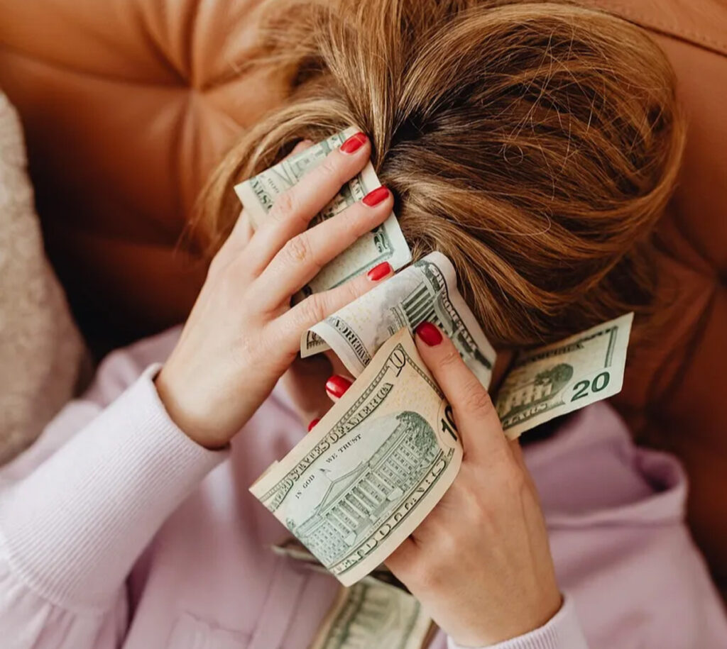 這張圖片展現了一位女性正面臨財務壓力，頭上放著鈔票，表達出她對經濟困難的焦慮。適合與財務壓力相關的內容或求助資源